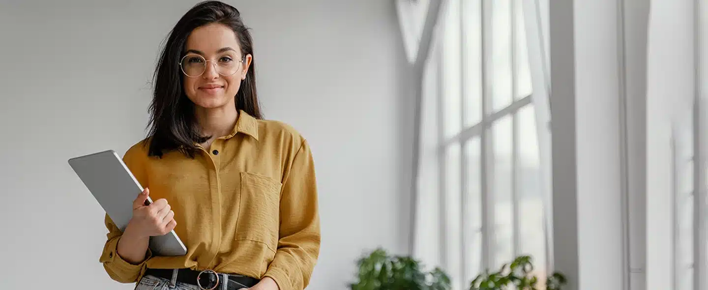 No lado esquerdo da imagem tem uma mulher jovem com camisa marrom, segurando um ipad. Atrás tem uma janela e cortinas brancas e logo abaixo tem algumas folhas verdes de plantas. Imagem usada para ilustrar o artigo sobre Mercado livre de energia vale a pena