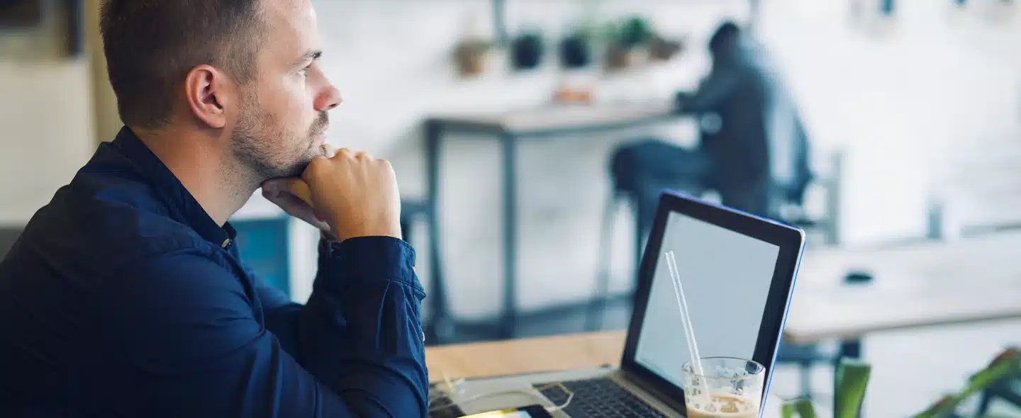 No lado esquerdo da imagem tem um homem com o queixo apoiando nas mãos fechadas em punho, pensando, em frente a um notebook aberto e um copo com um líquido e um canudo transparente. Ao fundo tem um homem sentado de costas e desfocado para exemplificar artigo sobre Placa solar ou Mercado Livre de energia: qual é a melhor opção?