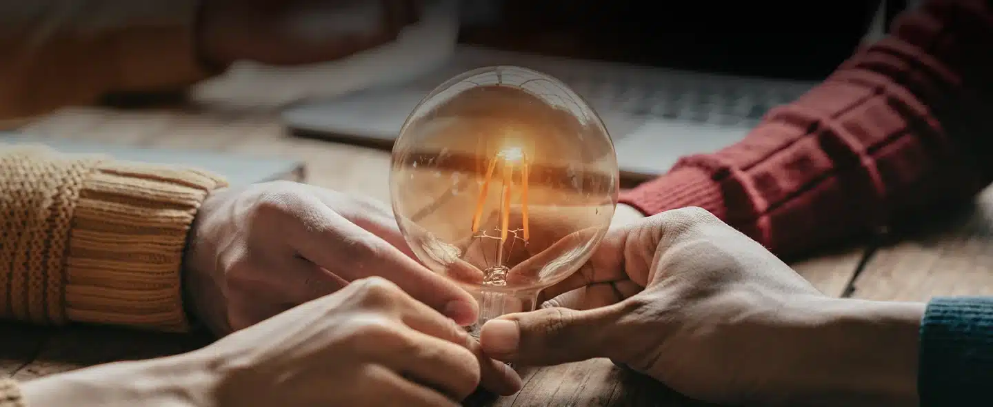 Quatro mãos segurando sob uma mesa segurando uma lampada que está emitndo uma luz amarela, quase laranja, para exemplificar o artigo sobre O que é uma cooperativa de energia renovável: como funciona, benefícios e quem pode participar