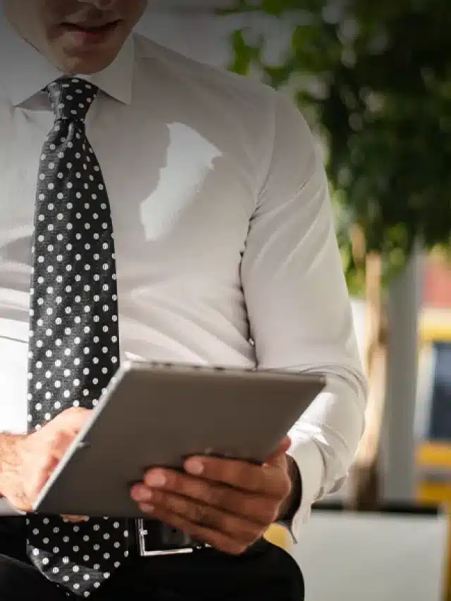 Um homem de camisa branca, gravata cinza semi sentado numa mensa segurando um ipad para exemplificar o artigo sobre "Empresa de energia solar: tudo que precisa saber antes de contratar uma"