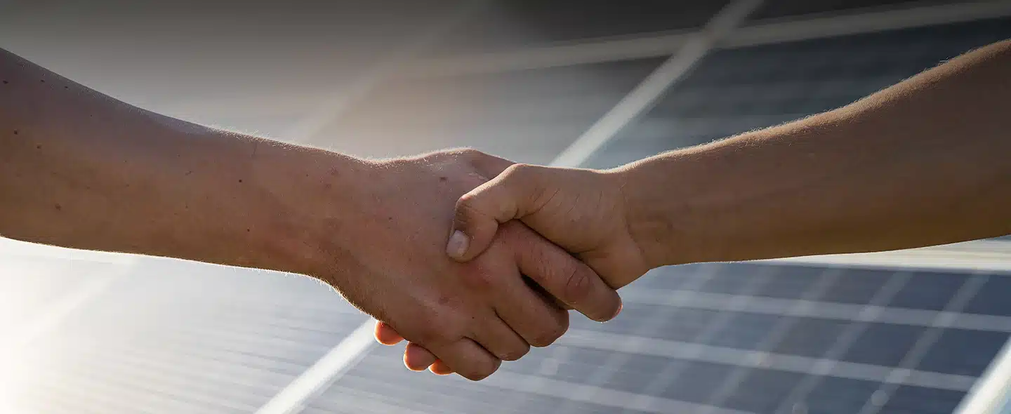 Na imagem tem duas mãos dando um aperto de mão e ao fundo um painel solar para exemplificar artigo sobre Descubra o que impacta no custo da energia solar