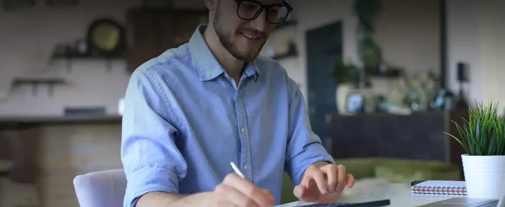 Homem jovem, de óculos, barba feita e camisa azul com mangas enroladas até a altura dos cotovelos. Ele está sorrindo segurando uma caneta para exemplificar artigo sobre como calcular custo de energia solar