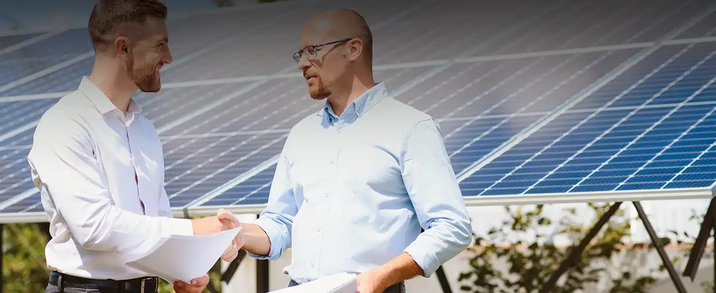 Dois homens se dando um aperto de mãos, logo atrás tem um painel solar desfocado para artigo sobre Aluguel de energia solar: como funciona?