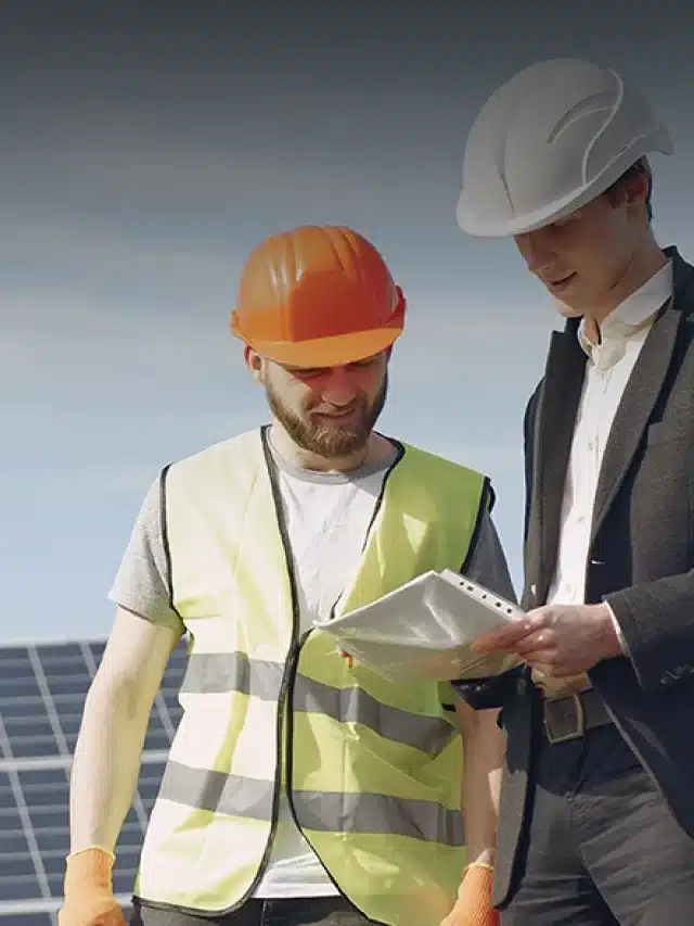 Na imagem, da direita para esquerda há dois homens com capacete. O da esquerda usa um coleto amarelo com faixas cinzas de API e outro, na direita, usa um terno. Ambos estão olhando para baixo, pois o homem de terno segura uma prancheta para exemplificar o artigo sobre Aprenda como entender a conta de luz com energia solar e dicas para economizar energia