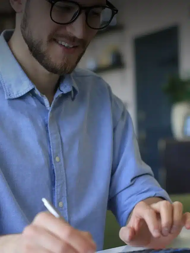 Homem jovem, de óculos, barba feita e camisa azul com mangas enroladas até a altura dos cotovelos. Ele está sorrindo segurando uma caneta para exemplificar artigo sobre como calcular custo de energia solar