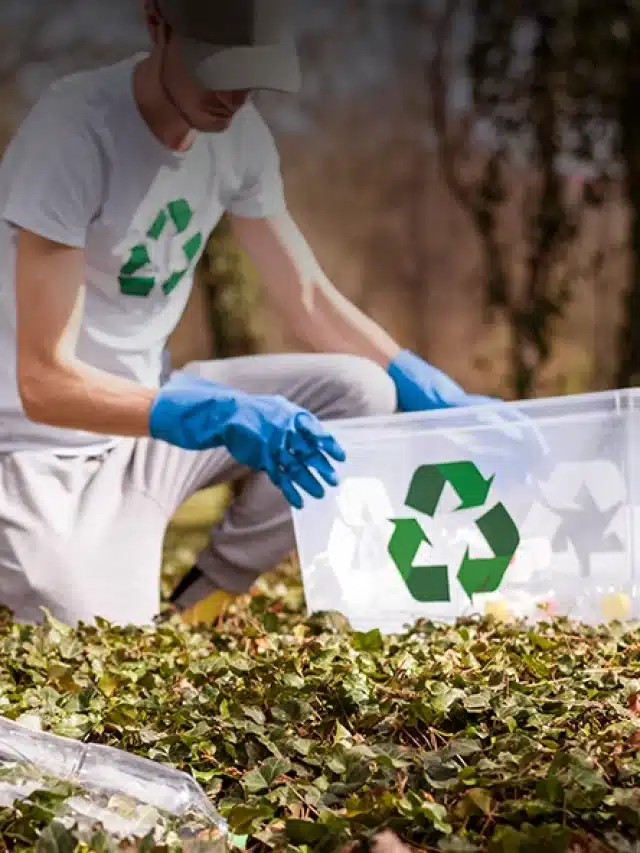 Rapaz com camisa com o simbolo de reciclável segura recipiente com o mesmo simbolo enquanto está abaixado no chão coletando garrafas pets para exemplificar artigo sobre Saiba o que é geração distribuída e como pode ajudar o meio ambiente