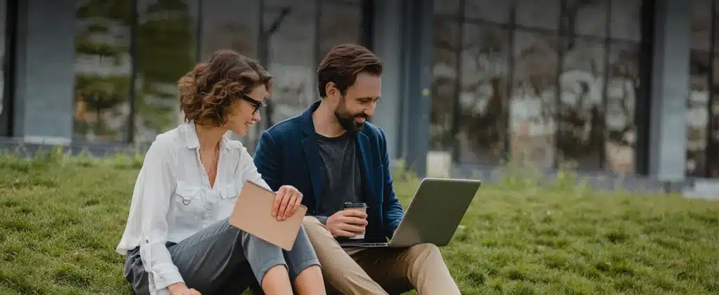 Homem e mulher sentados na grama olhando o laptop enquanto sorriem para artigo sobre As vantagens das tecnologias sustentáveis no Brasil