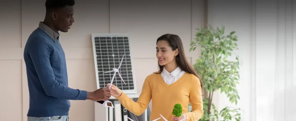 Homem e mulher seguram uma réplica de uma turbina eólica e logo atrás há uma placa solar para artigo sobre Descubra o que é energia compartilhada e como ela funciona