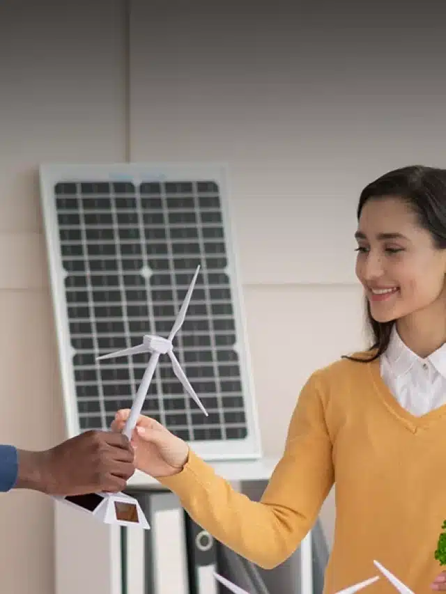 Homem e mulher seguram uma réplica de uma turbina eólica e logo atrás há uma placa solar para artigo sobre Descubra o que é energia compartilhada e como ela funciona