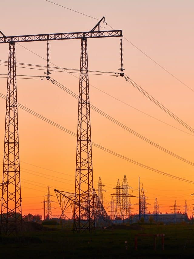 High voltage electricity towers in field at sunset and clear blue sky. Dark silhouettes of repeating power lines on orange sunrise. Electricity generation, transmission, and distribution network