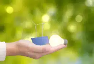Mão segurando lâmpada acesa, painel solar e turbinas eólicas, simbolizando energia acessível e limpa para um futuro sustentável.