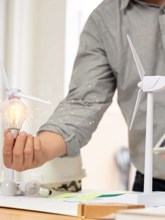 close-em-uma-lampada-um-engenheiro-esta-sentado-segurando-uma-lampada-e-um-modelo-de-turbina-eolica-com-paineis-solares-e-uma-casa-modelo-sobre-a-mesa