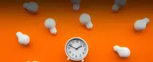 Relógio e lâmpadas brancas no fundo laranja para Saiba qual o melhor horário para consumir energia