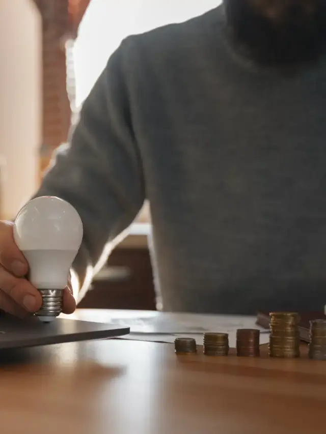 Homem segurando uma lâmpada LED enquanto calcula finanças e economiza dinheiro com energia por assinatura em casa, ao lado de pilhas de moedas e notebook.