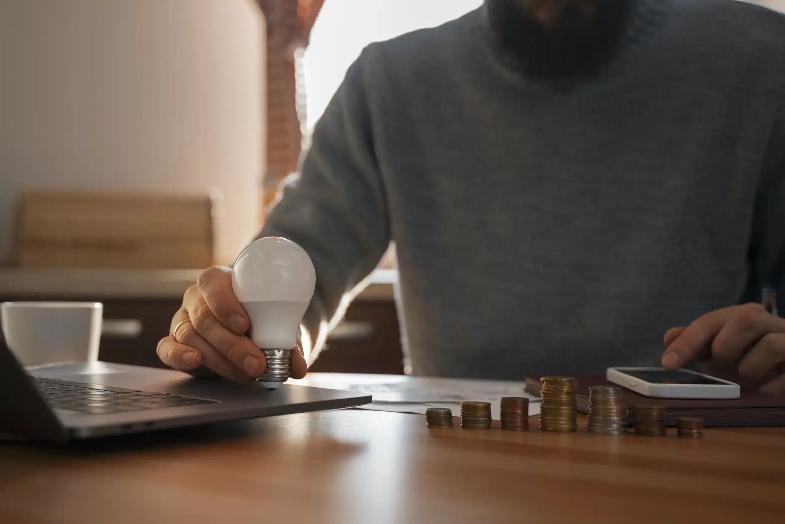 Homem segurando uma lâmpada LED enquanto calcula finanças e economiza dinheiro com energia por assinatura em casa, ao lado de pilhas de moedas e notebook.
