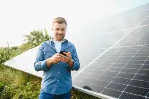 Homem sorridente usando celular em frente a painéis solares, representando a importância do pós-venda em usinas solares para garantir satisfação e relacionamento com o cliente.