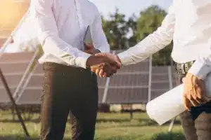 Empresários selando parceria com aperto de mãos em frente a usinas solares, representando o desafio da captação de clientes para usinas solares e como superá-lo.
