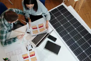 Dois profissionais analisando projetos arquitetônicos e modelos de energia solar ao lado de um painel fotovoltaico, com apoio de um software de gestão para usinas solares.