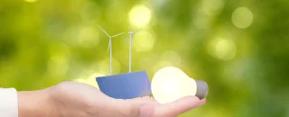 Mão segurando lâmpada acesa, painel solar e turbinas eólicas, simbolizando energia acessível e limpa para um futuro sustentável.