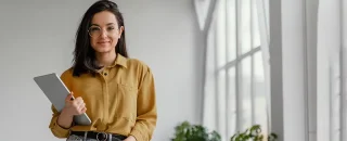 No lado esquerdo da imagem tem uma mulher jovem com camisa marrom, segurando um ipad. Atrás tem uma janela e cortinas brancas e logo abaixo tem algumas folhas verdes de plantas. Imagem usada para ilustrar o artigo sobre Mercado livre de energia vale a pena