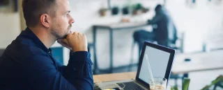 No lado esquerdo da imagem tem um homem com o queixo apoiando nas mãos fechadas em punho, pensando, em frente a um notebook aberto e um copo com um líquido e um canudo transparente. Ao fundo tem um homem sentado de costas e desfocado para exemplificar artigo sobre Placa solar ou Mercado Livre de energia: qual é a melhor opção?