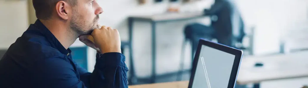 No lado esquerdo da imagem tem um homem com o queixo apoiando nas mãos fechadas em punho, pensando, em frente a um notebook aberto e um copo com um líquido e um canudo transparente. Ao fundo tem um homem sentado de costas e desfocado para exemplificar artigo sobre Placa solar ou Mercado Livre de energia: qual é a melhor opção?