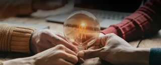 Quatro mãos segurando sob uma mesa segurando uma lampada que está emitndo uma luz amarela, quase laranja, para exemplificar o artigo sobre O que é uma cooperativa de energia renovável: como funciona, benefícios e quem pode participar