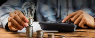 Duas mãos masculinas segurando uma lampada apagada, sob uma colune de moedas e seguem mais três pequenas colunas de moedas em ordem decrescente de tamanho. Na imagem também tem uma calculadora e um caderno de folhas brancas. Ao fundo aparece uma parte da camisa azul do homem levemente desfocado para exemplificar o artigo sobre sistema de energia solar para empresas
