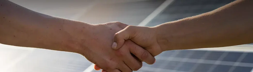 Na imagem tem duas mãos dando um aperto de mão e ao fundo um painel solar para exemplificar artigo sobre Descubra o que impacta no custo da energia solar