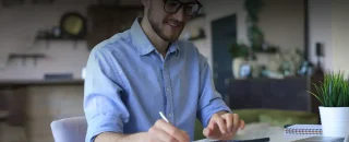 Homem jovem, de óculos, barba feita e camisa azul com mangas enroladas até a altura dos cotovelos. Ele está sorrindo segurando uma caneta para exemplificar artigo sobre como calcular custo de energia solar