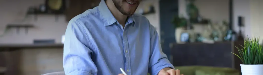 Homem jovem, de óculos, barba feita e camisa azul com mangas enroladas até a altura dos cotovelos. Ele está sorrindo segurando uma caneta para exemplificar artigo sobre como calcular custo de energia solar