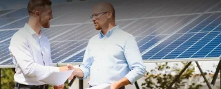 Dois homens se dando um aperto de mãos, logo atrás tem um painel solar desfocado para artigo sobre Aluguel de energia solar: como funciona?