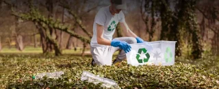 Rapaz com camisa com o simbolo de reciclável segura recipiente com o mesmo simbolo enquanto está abaixado no chão coletando garrafas pets para exemplificar artigo sobre Saiba o que é geração distribuída e como pode ajudar o meio ambiente