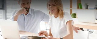 Homem e mulher com roupa branca em frente um laptop branco enquanto usam e olhem uma prancheta com atenção para artigo de 5 formas de economizar energia