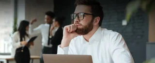 Homem jovem de óculos com armação preta com blusa branca de frente para laptop com olhar distante para artigo sobre Descubra se precisa de painel solar para ter energia solar por assinatura