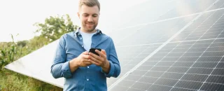 Homem sorridente usando celular em frente a painéis solares, representando a importância do pós-venda em usinas solares para garantir satisfação e relacionamento com o cliente.