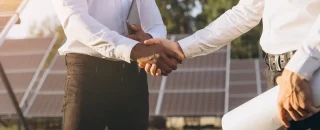 Empresários selando parceria com aperto de mãos em frente a usinas solares, representando o desafio da captação de clientes para usinas solares e como superá-lo.
