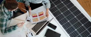 Dois profissionais analisando projetos arquitetônicos e modelos de energia solar ao lado de um painel fotovoltaico, com apoio de um software de gestão para usinas solares.