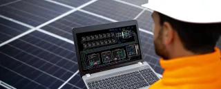 Engenheiro analisando dados em laptop sobre o desempenho de uma plataforma de gestão para usinas solares, em frente a painéis fotovoltaicos.