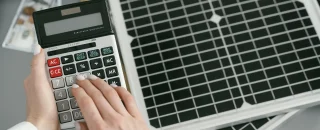 Pessoa calculando custos ao lado de painéis solares, simbolizando o investimento em uma micro usina solar e o retorno financeiro com energia limpa.