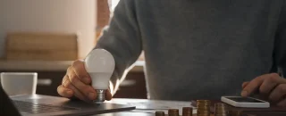 Homem calculando finanças com moedas sobre a mesa e segurando uma lâmpada, simbolizando a escolha de economizar com energia por assinatura usando tecnologia e planejamento financeiro.